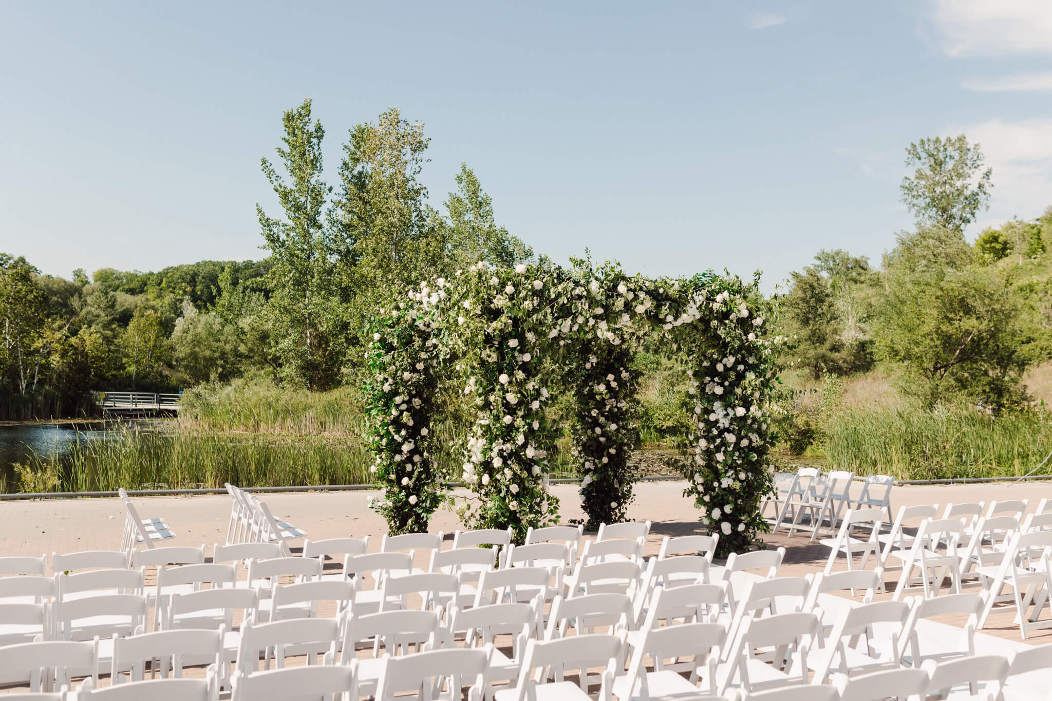 Evergreen Brick Works outdoor summer wedding setup along pond and green trees. Empty white chairs and two white rose and greenery covered arbours at the end of the aisle.