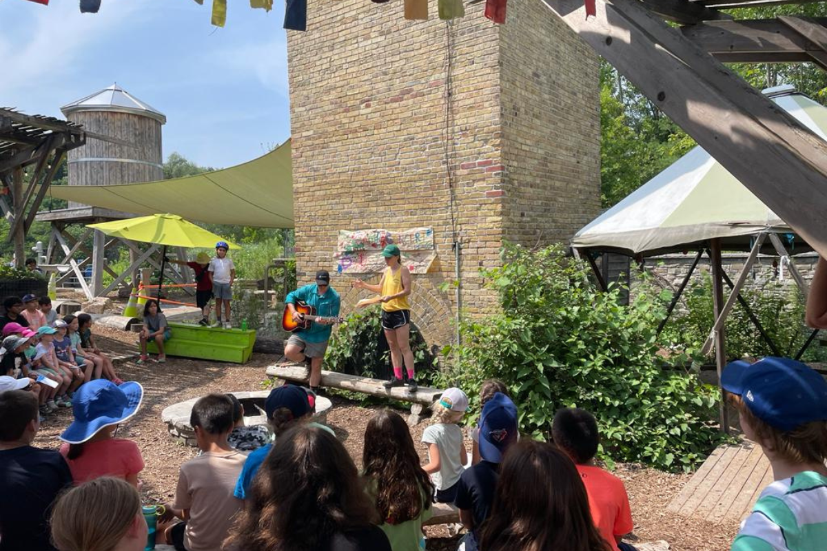 Two artists performing, one with an acoustic guitar and the other singing with some papers, tp a group of children in front of an unlit fire pit, brick chimney in the summer.