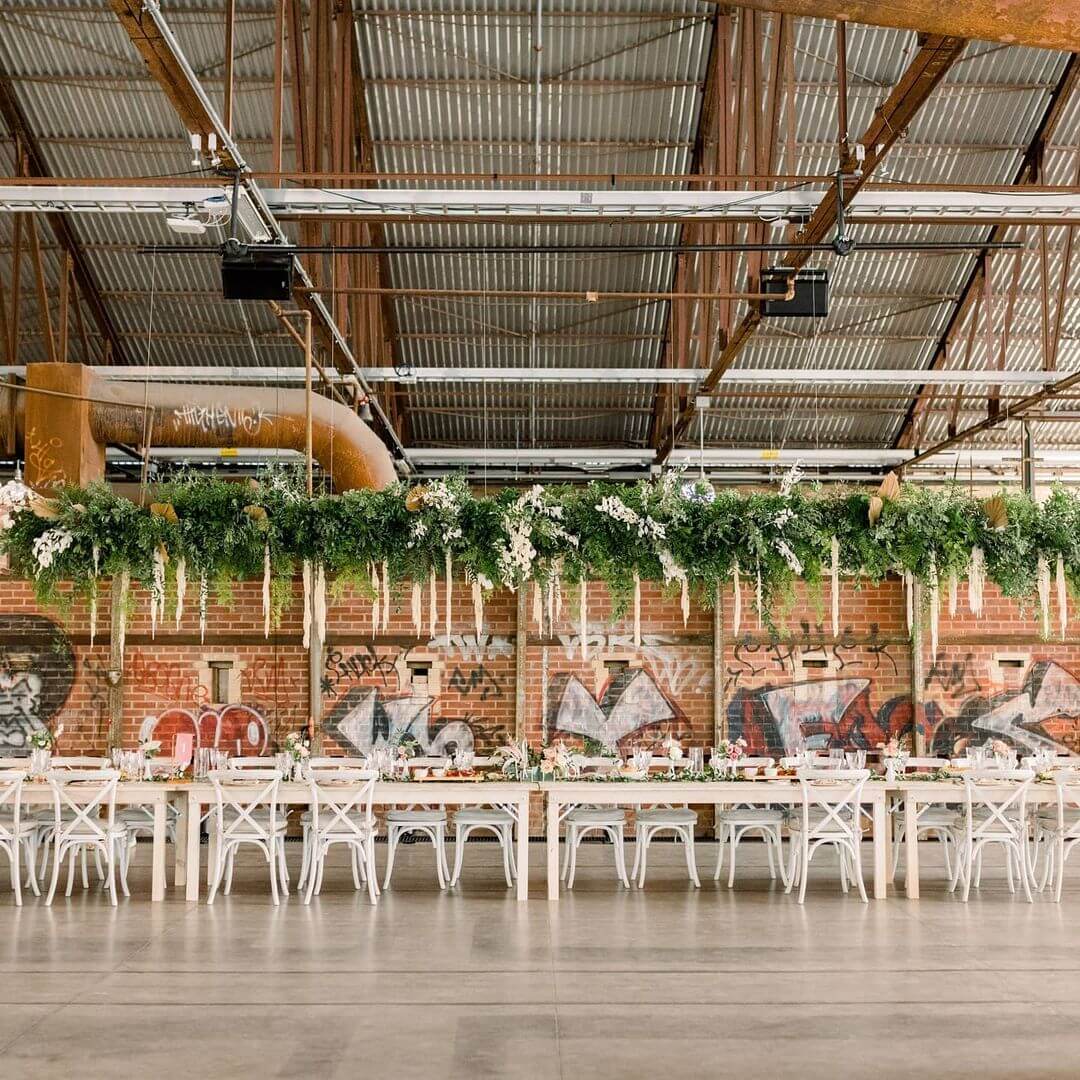 Evergreen Brickworks CRH wedding setup with long white tables and chairs, green floral arrangement hanging along the ceiling above the tables and exposed brick with graffiti backdrop.