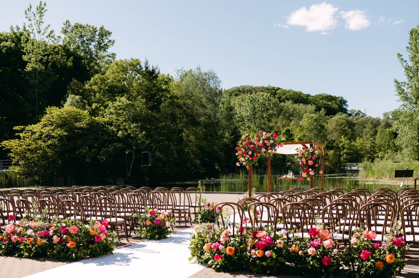 Evergreen Brick Works summer outdoor wedding setup with empty wooden chairs and bright florals, pond, trees and blue sky backdrop.