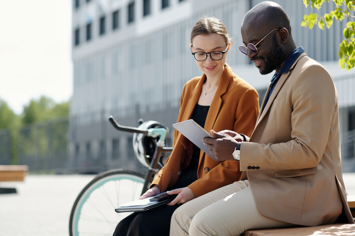Two people sit on bench outside in city hold and looking at tablet