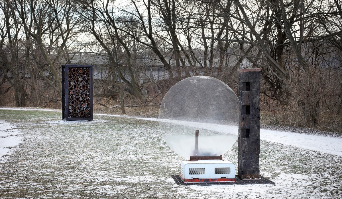 Virginia Overton art sculpture using industrial and natural looking recycled materials outside on grass with snow on the ground and winter trees. Round plastic item on a stand beside wooden vertical beam a few yards away from metal framed circular items.