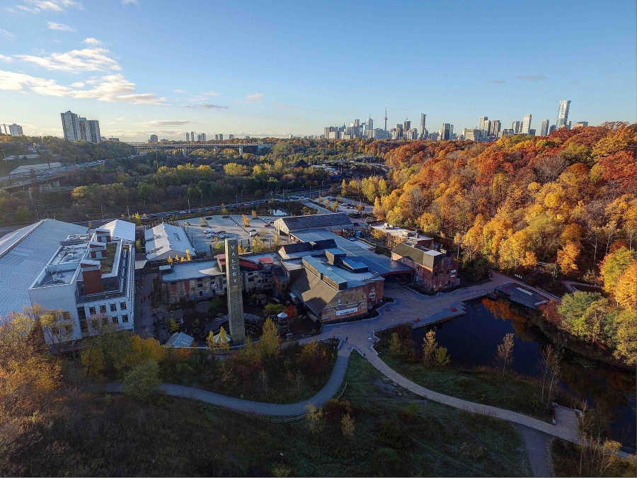 Evergreen Brick works drone shot. Collection of building with pathways leading into fall coloured forest and the City of Toronto skyline.