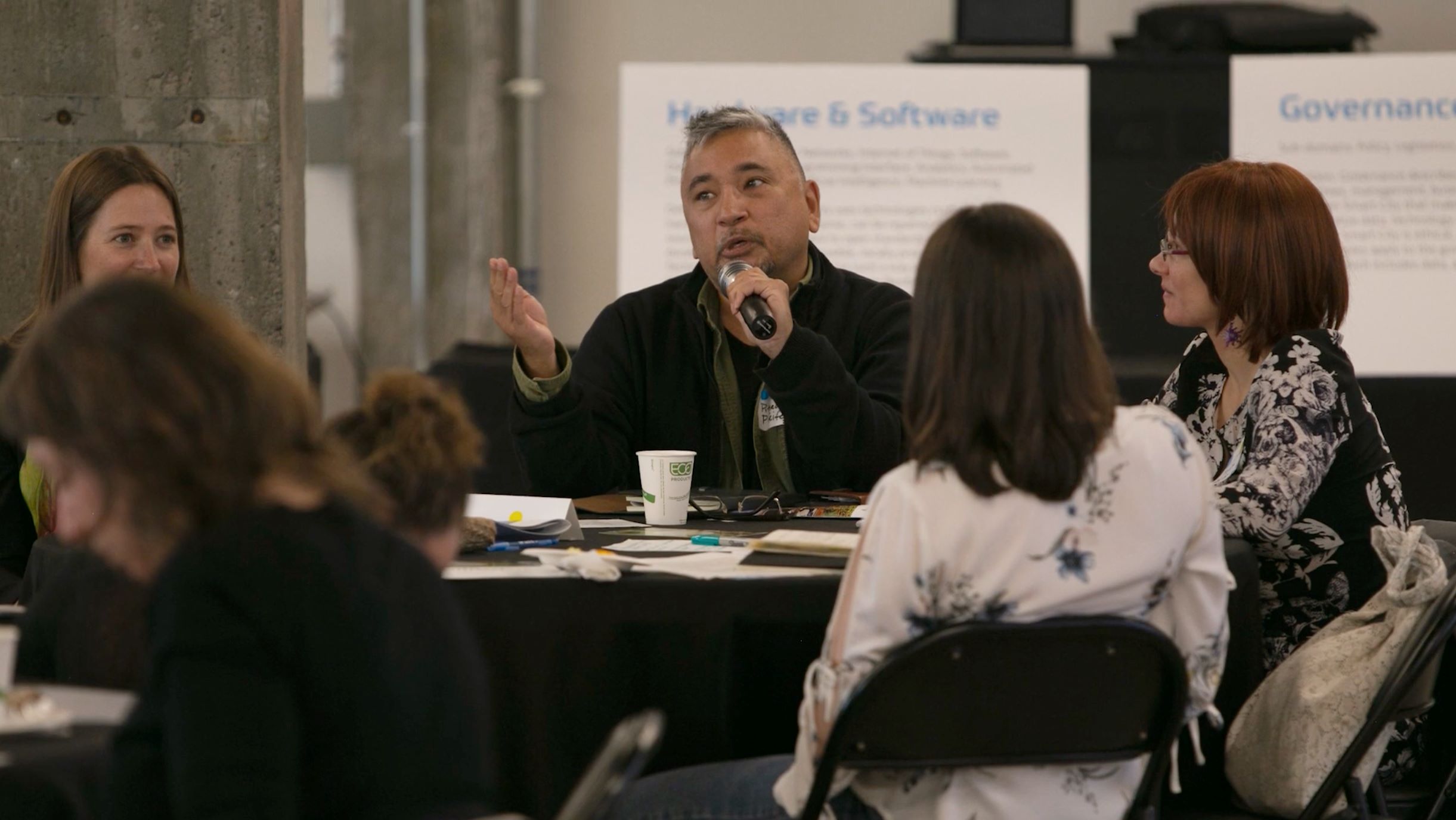 Man speaking into microphone while seated at a conference table with other people