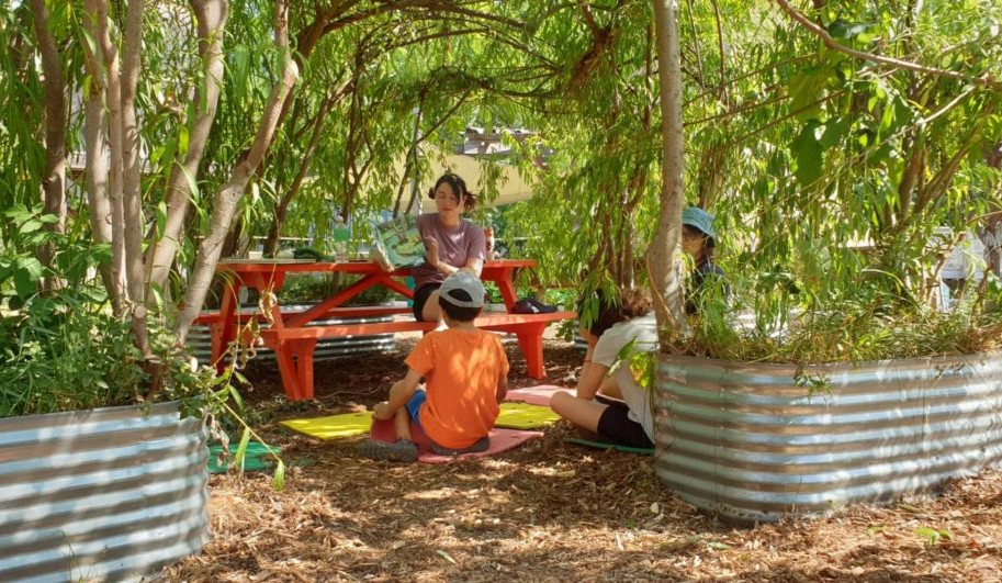 Children sit on the ground while an older youth reads them a book from a picnic table in a shady garden