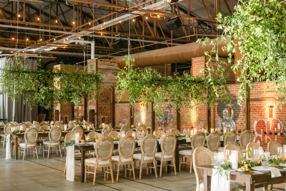 Event space with exposed brick and metal beams on ceiling. Wicker backed chairs and tables decorated for a wedding with floral greenery hanging.