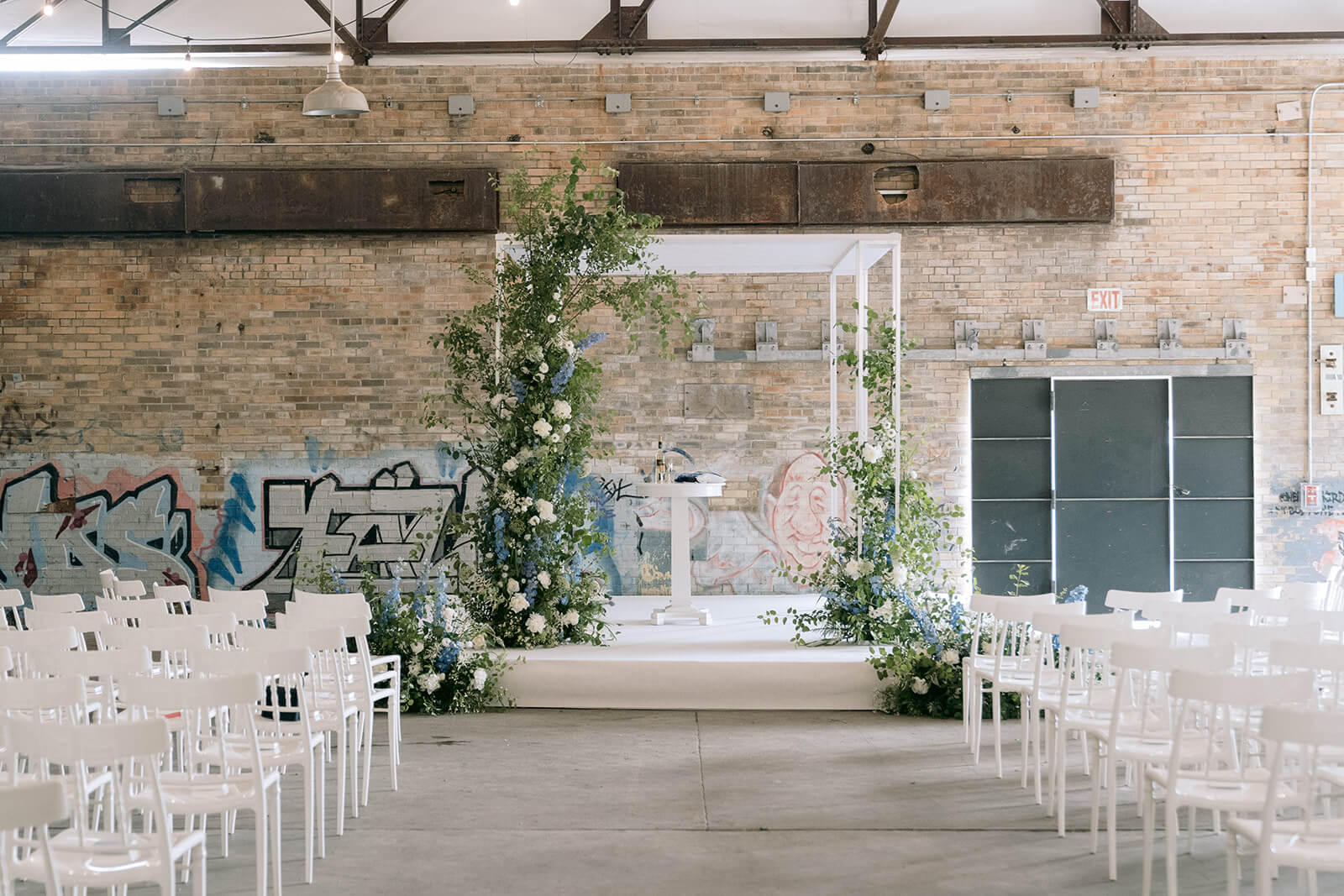 Evergreen Brick Works Koerner Gardens white stage and arbour with green and white florals. White empty chairs along the aisle with exposed brick and graffiti backdrop.