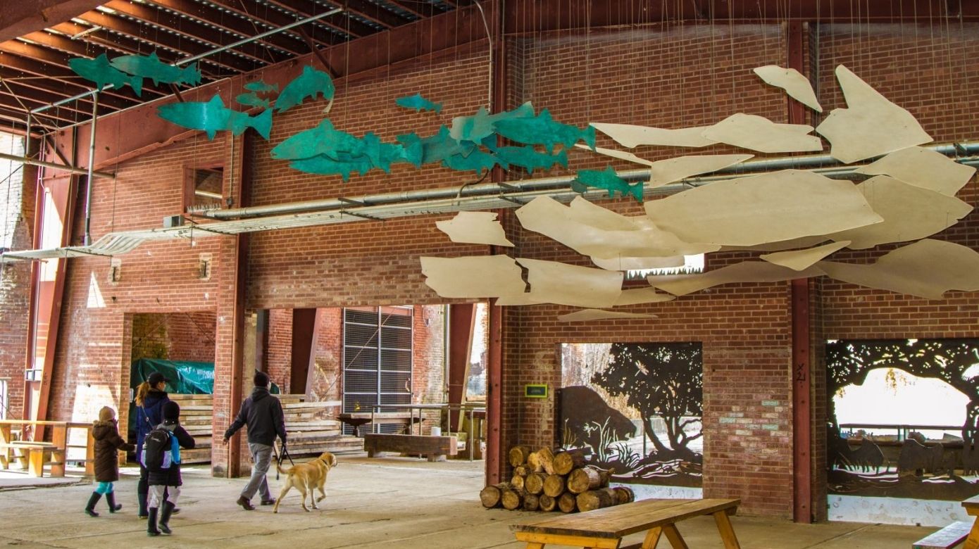 Evergreen Brick Works Pavilion with exposed red brick, a family walking their yellow Labrador retriever and a hanging art structure of teal metal fish cutouts and other beige shapes.