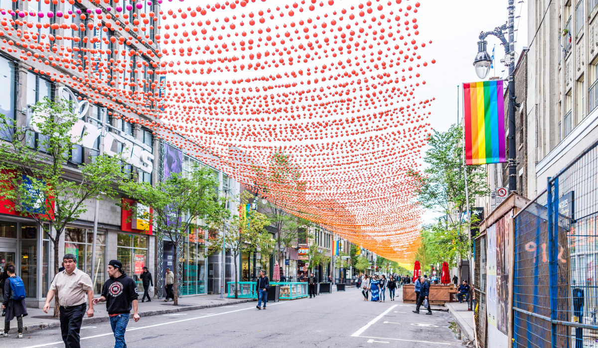 Pride flag hung from light post and pedestrians walking through a closed city street decorated with hundreds of coloured balls creating an ombré effect for pride.
