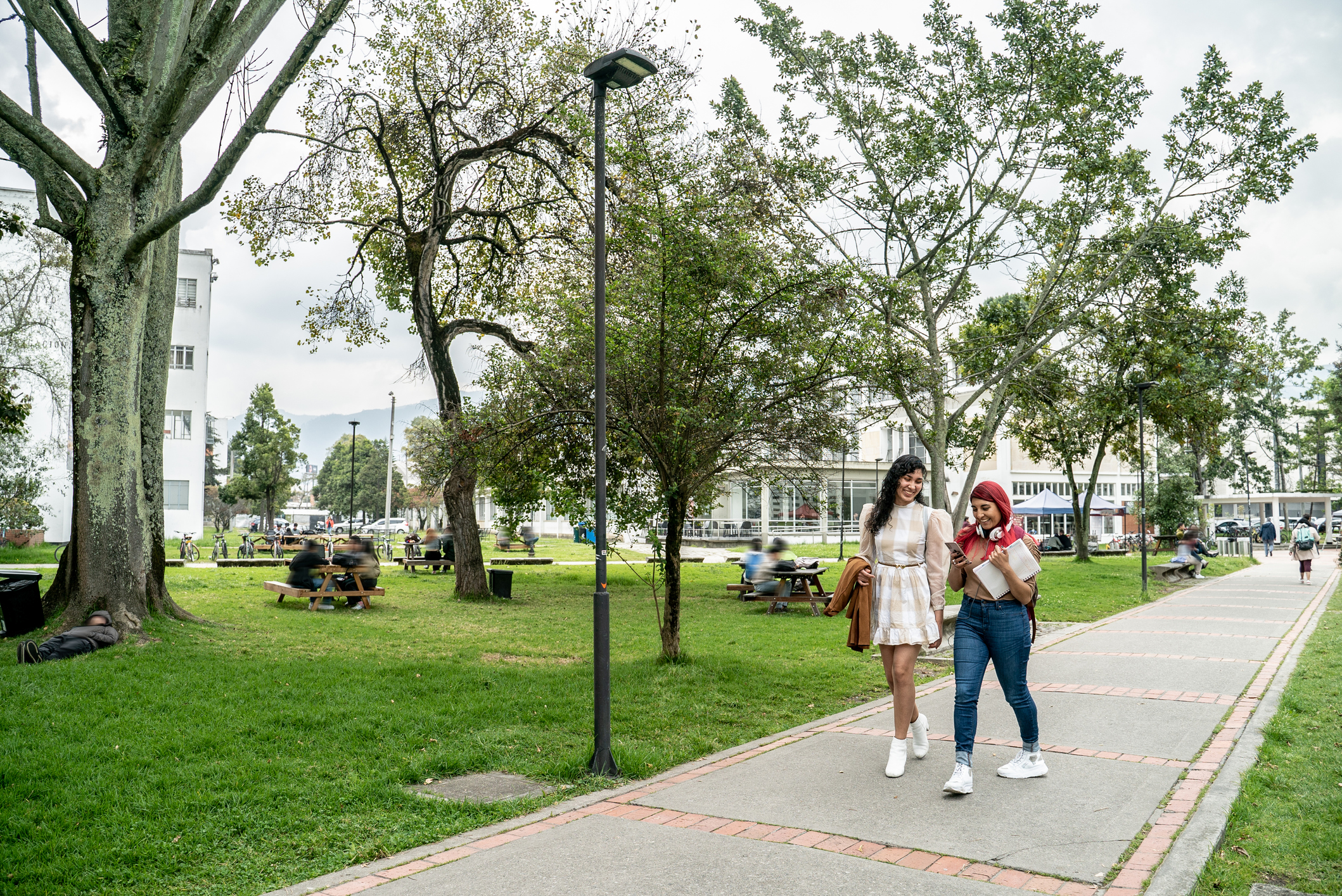 Two young women walking in a public park in a town with picnic tables and other individuals blurred in the background.