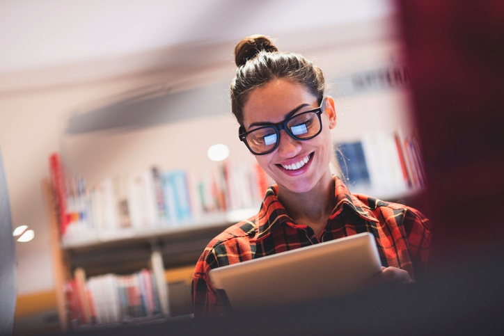 Young person with glasses looking up resources on a tablet, screen shows in glasses giving off a reflection. Books in background
