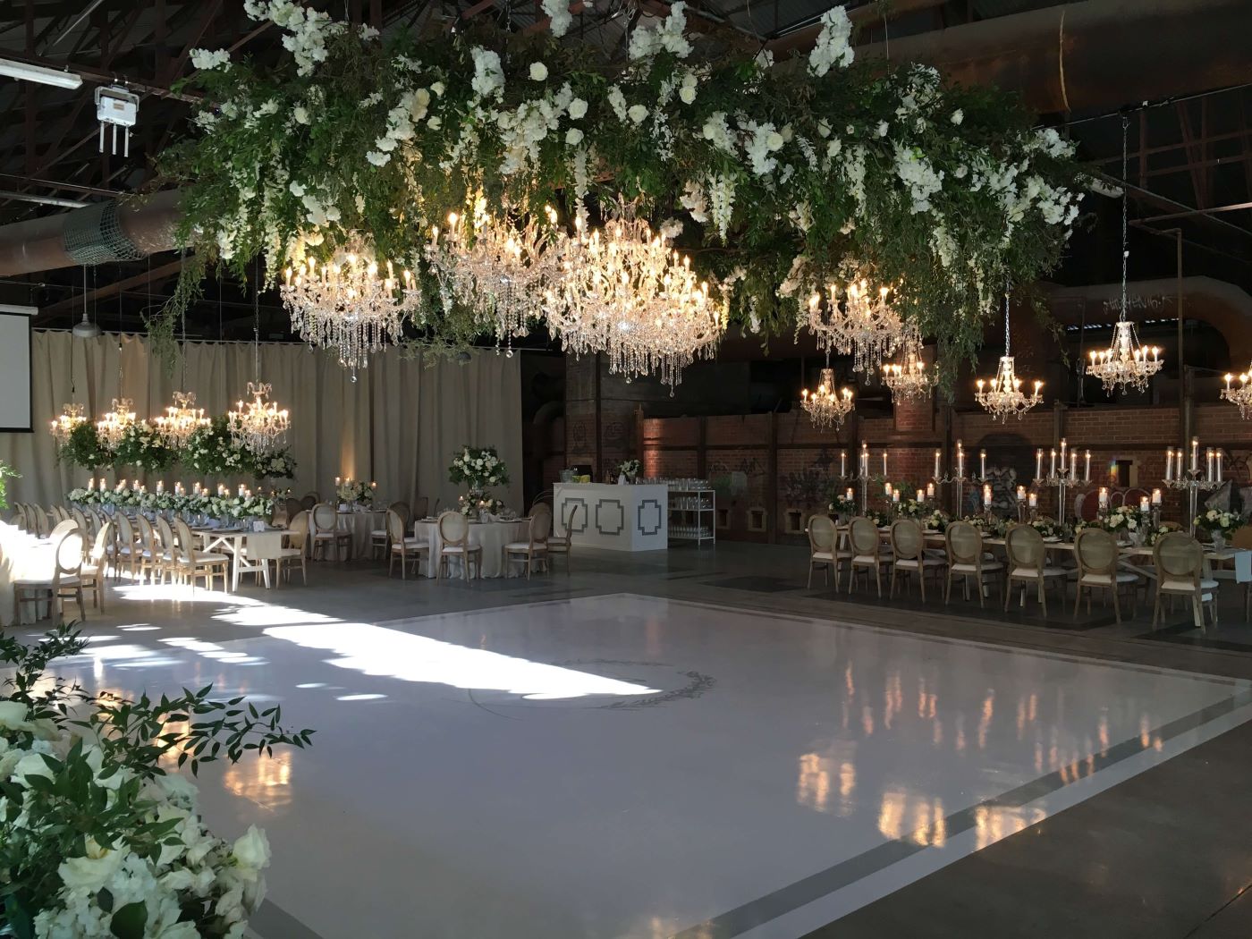 Evergreen Brickworks CRH Gallery wedding setup with crystal chandeliers lit up and covered in white roses and greenery. Large white dance floor in the middle of wedding tables.