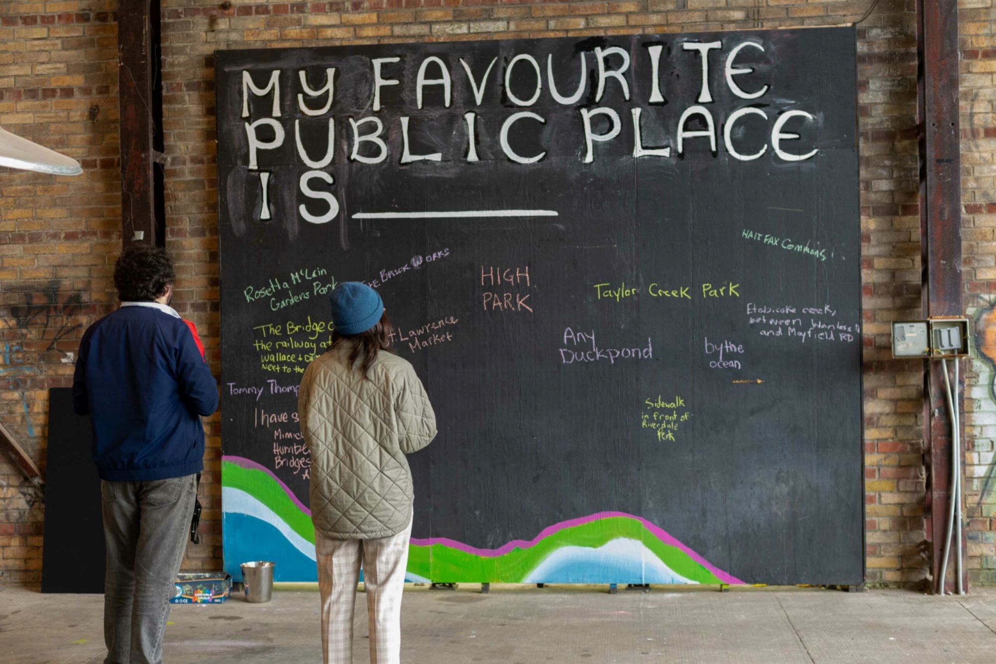 People stand in front of a chalkboard where people wrote examples of their favourite public places.