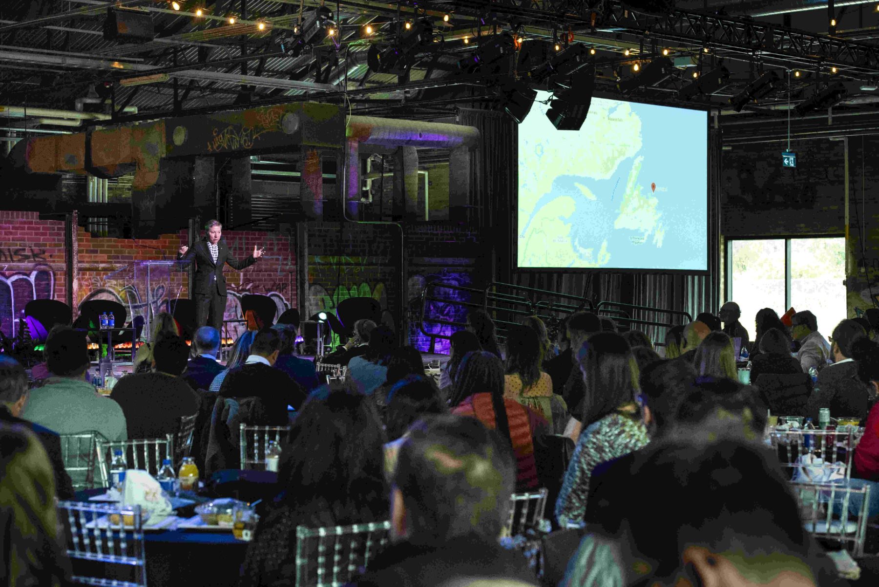people sit at tables at conference listening to speaker on stage
