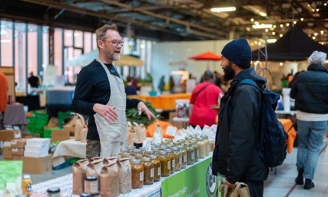A visitor at a farmers market speaks with a friendly market vendor