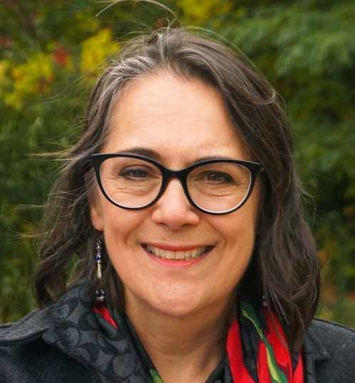 Sheila Boudreau has shoulder length brown hair, brown eyes and is wearing black rimmed eyeglasses and a colourful scarf.