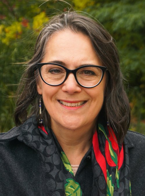 Head shot of Sheila Boudreau smiling wearing black-rimmed glasses and a colourful scarf. She is outdoors with greenery in the background.
