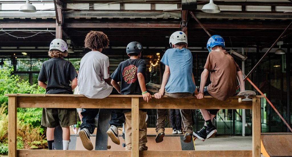 five young people sit on railing wearing helmets watching other young people skateboard