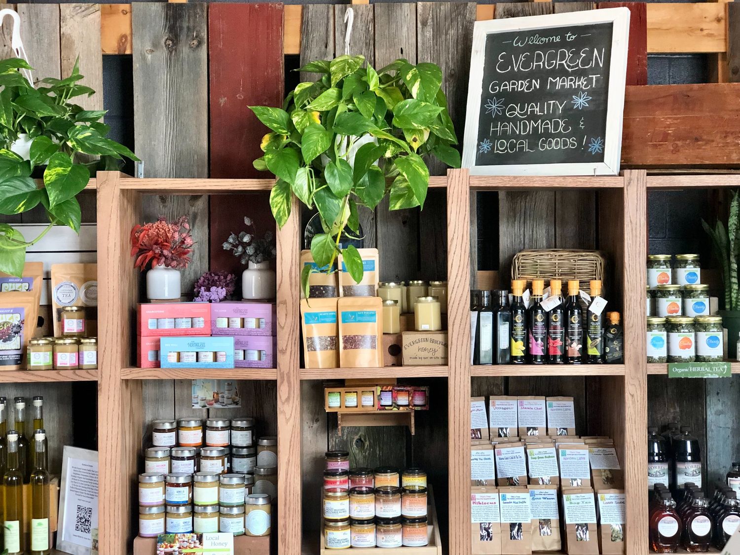 Variety of plants and products on a wooden shelf with a sign that reads 'Welcome to Evergreen Garden Market: quality handmade local goods