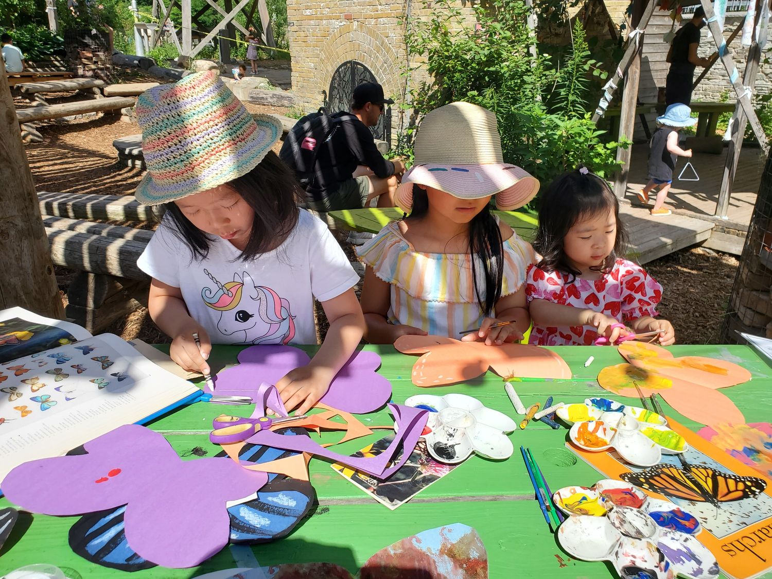 Three young children make butterfly crafts out of colourful construction paper and paint outdoors