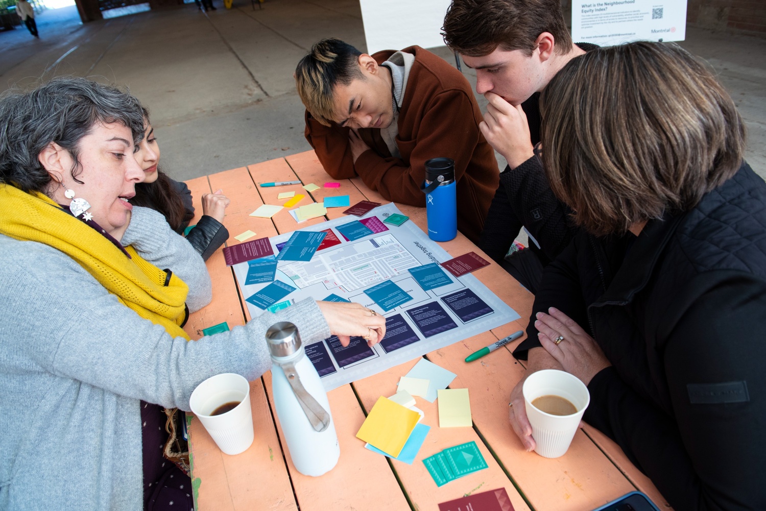 Five people sit at an orange picnic table with coffees and water while they play a boardgame