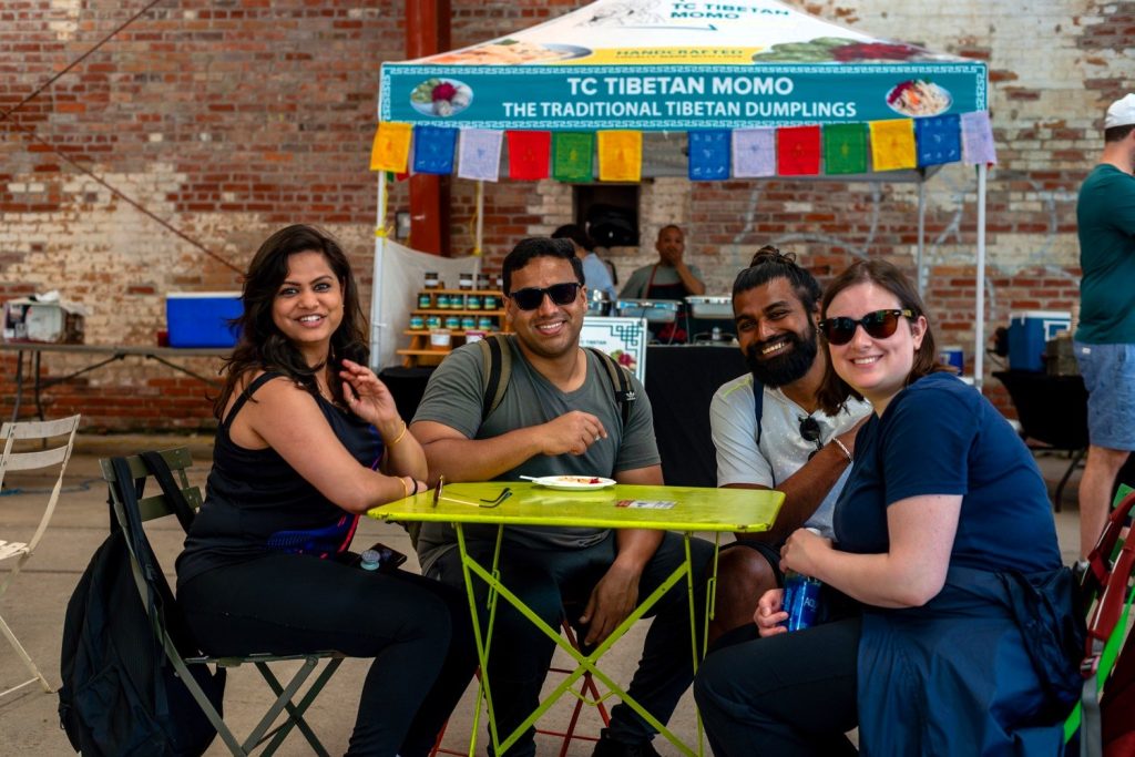 Four adults sit at a bright green table outdoors smiling in front of a food vendor