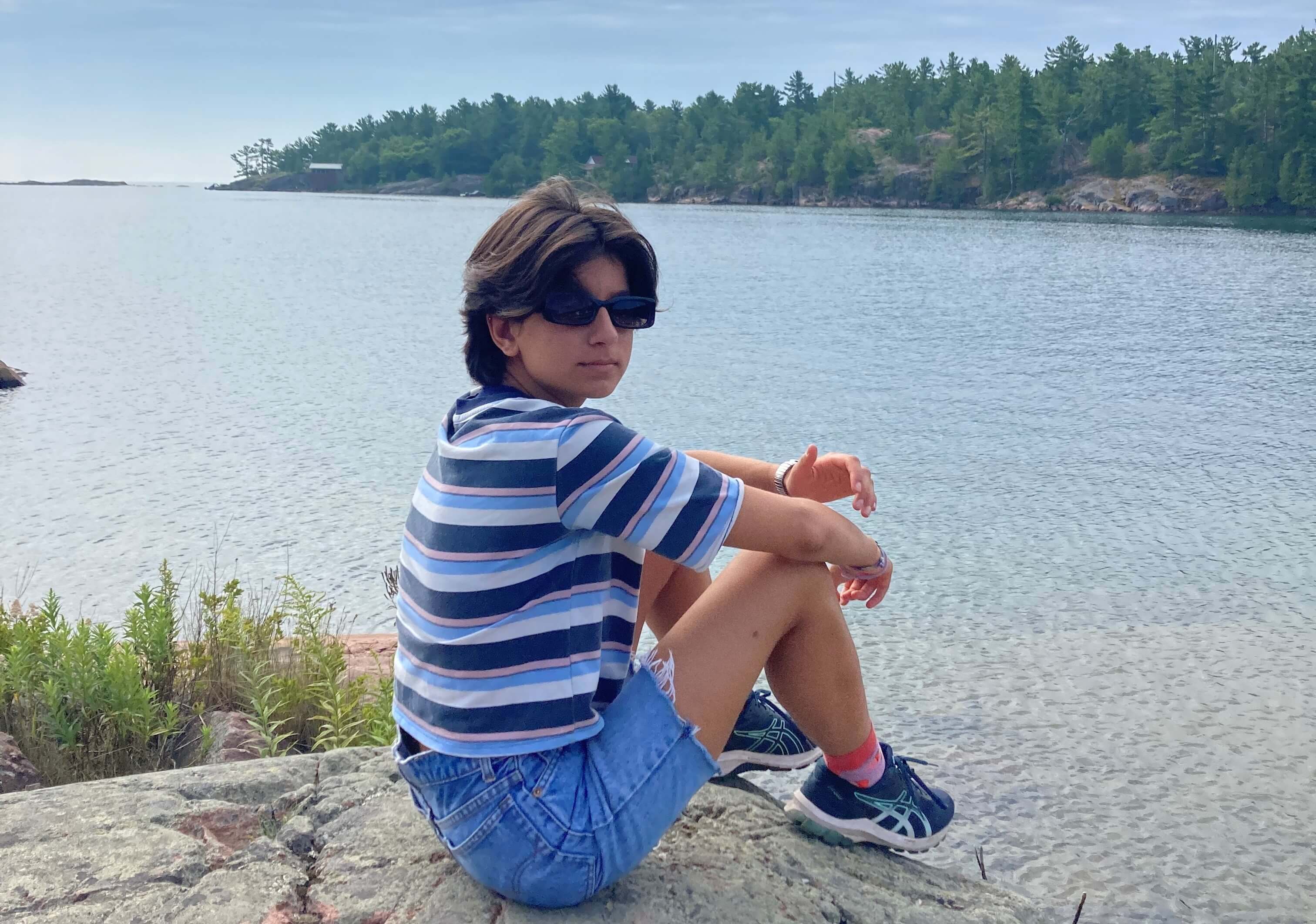 Anisa sits on a rock overlooking a lake with forest beyond the lake under a blue sky 