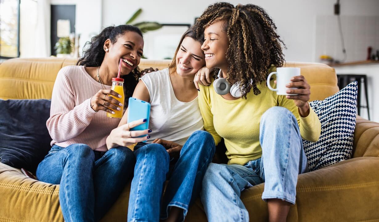 three women sit on a couch talking and laughing looking at phone